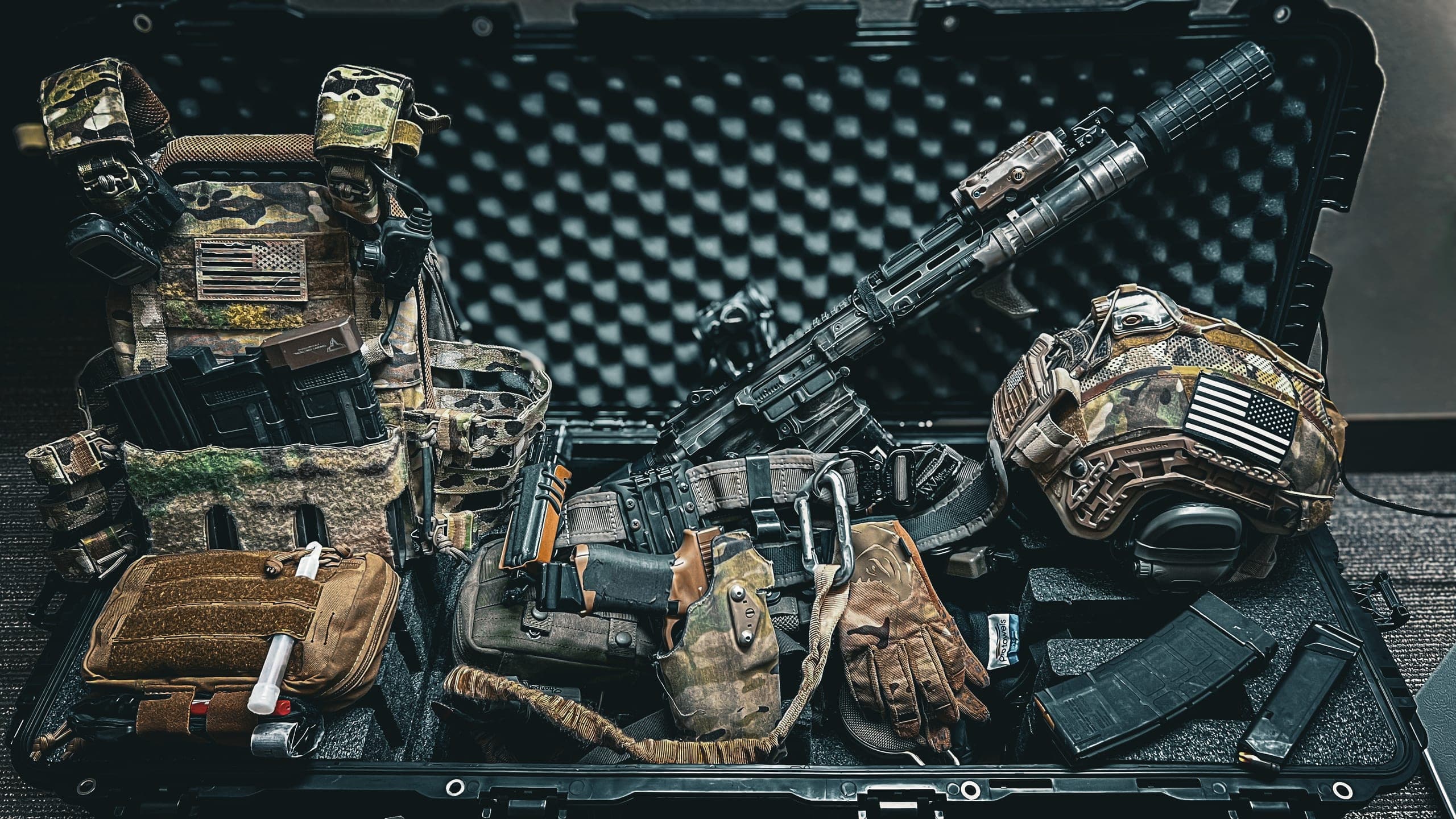 An open hard case packed with a plate carrier, a suppressed carbine, a helmet, a holster and a medical kit