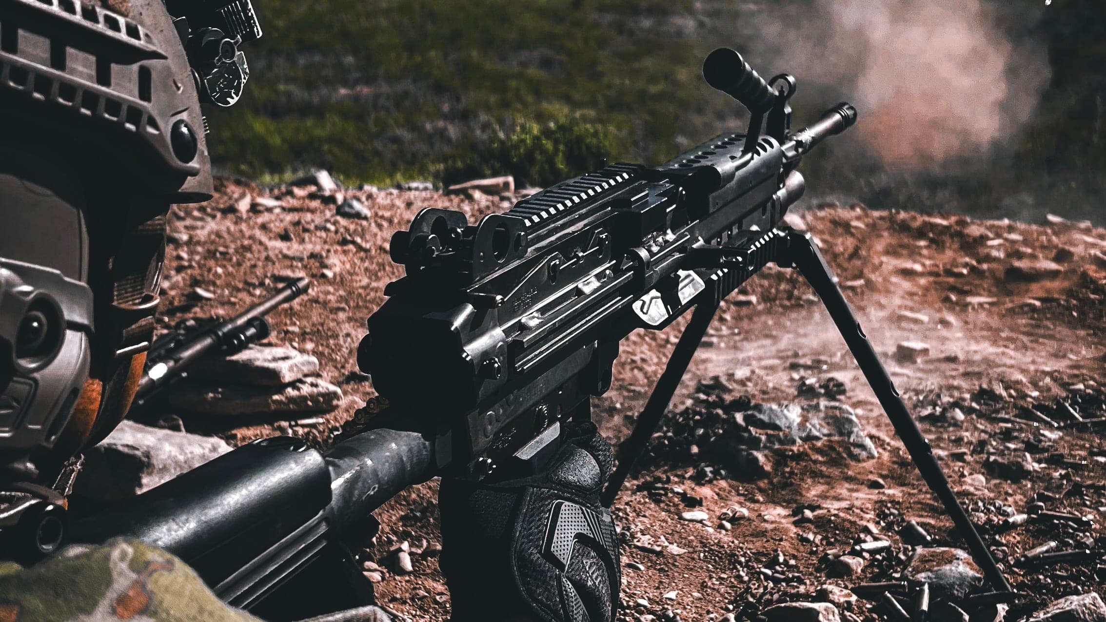 A belt-fed machine gun on its bipod, firing on a rocky range
