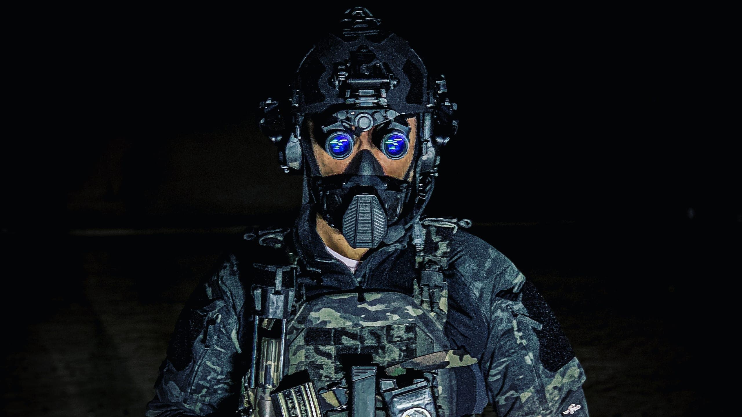 An operator in a gas mask and night vision with a loaded plate carrier, against black