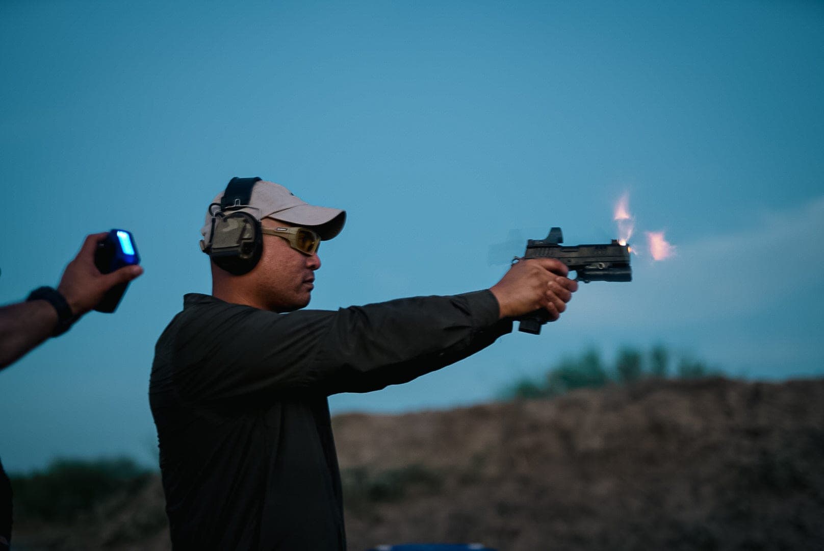 A shooter firing a red-dot pistol at dusk, muzzle flash lit against a blue sky, an instructor timing the string beside him