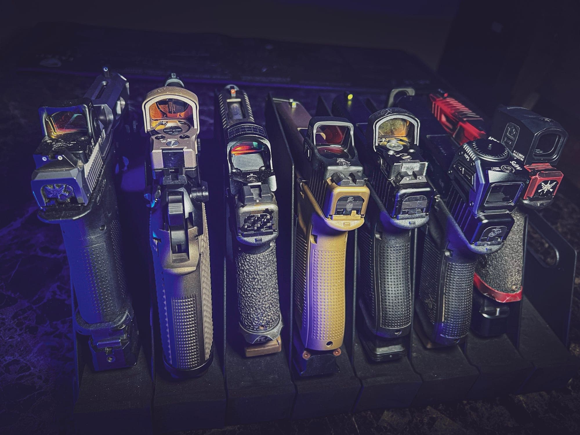 Seven pistols standing muzzle-up in a rack, each wearing a different red dot optic